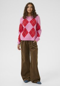 Göndör hajú nő rózsaszín és piros argyle mintás pulóverben, széles barna kordnadrágban és kék balettcipőben áll, egyik kezét a zsebében tartva.