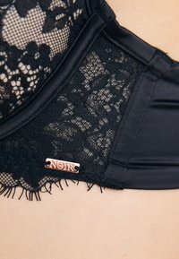 Svart blonde-bh med intrikate blomstermønstre, satindetaljer og et lite rosegull "NOIR"-merke på siden.