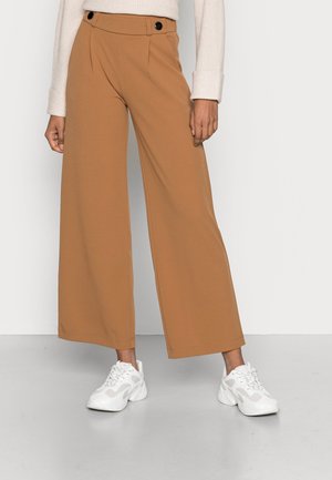 Osoba w szerokich spodniach camel i białych masywnych sneakersach stoi na tle jednolitego jasnego tła.