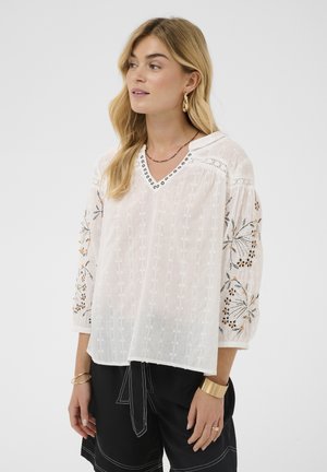 Blonde vrouw die een witte blouse draagt met geborduurde bloemenpatronen op de mouwen en zwarte broek met witte stiksels.