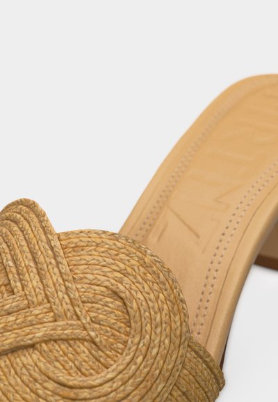 Közelkép egy világosbarna színű, fonott szalma muleszandálról, amelynek felsőrészén fonott csomó díszítés látható, és varrott talpa van.