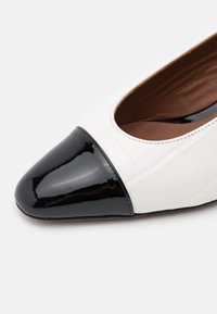 Bicolorní plochá obuv z bílé texturované kůže s lesklou černou špičkou. Viditelné švy podél otvoru.