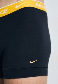 Boxers Nike negros con una cinturilla elástica amarilla que presenta el logotipo en blanco "NIKE" y un pequeño logo amarillo en la pierna.