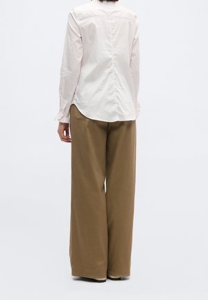 Personne portant un chemisier rose clair à manches longues et un pantalon marron à jambes larges, debout de dos face au spectateur sur un fond uni.