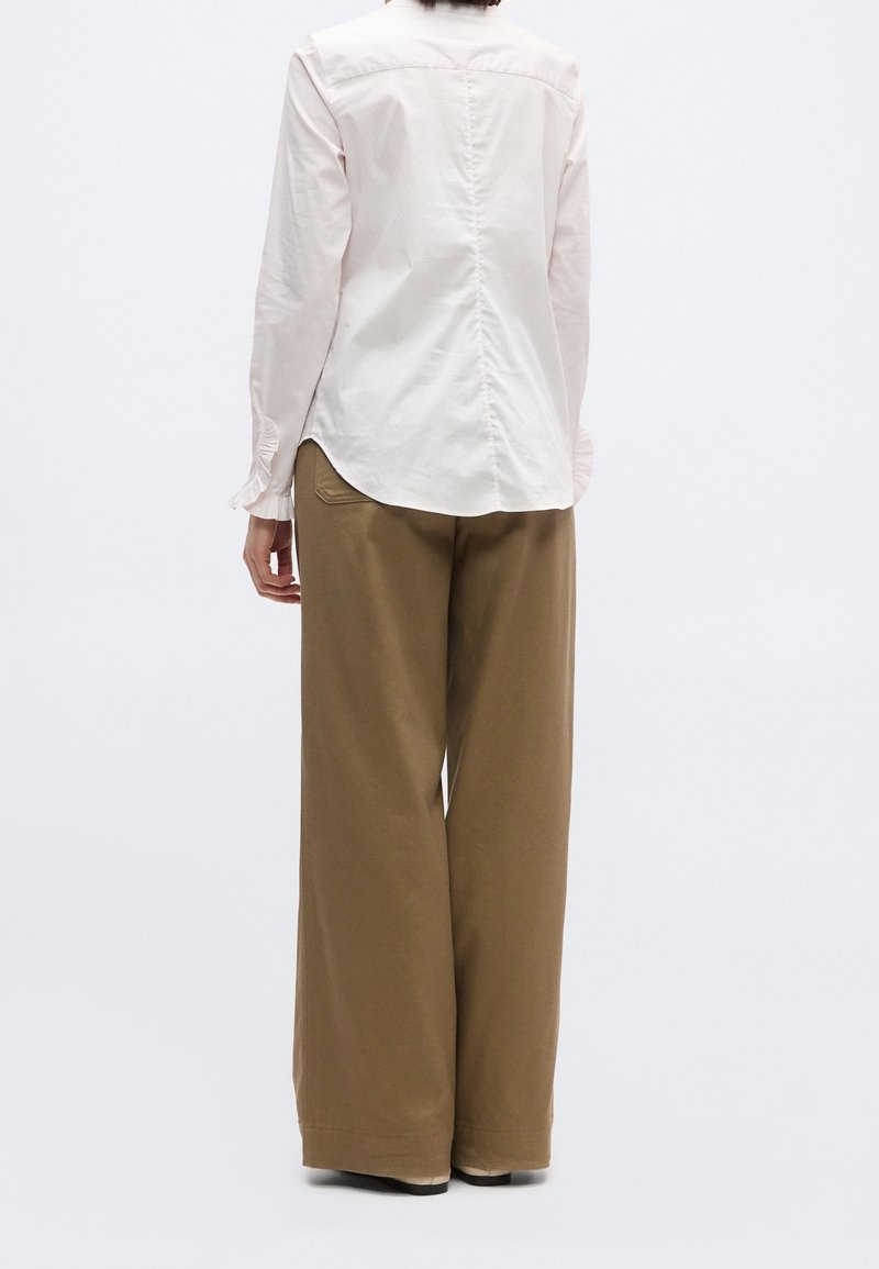 Personne portant un chemisier rose clair à manches longues et un pantalon marron à jambes larges, debout de dos face au spectateur sur un fond uni.