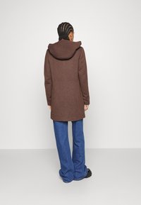 Brown wool coat featuring a large hood, knee-length cut, and loose fit, paired with wide-legged blue denim jeans.