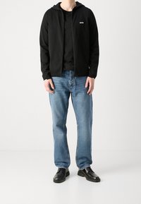Schwarze Zip-Up-Hoodie mit Logo, über einem schwarzen T-Shirt getragen, kombiniert mit blauen Jeans und schwarzen Schuhen. Minimalistisches und lässiges Outfit.
