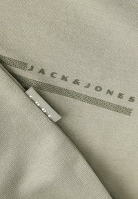 Μπεζ πλεκτό ύφασμα με τυπωμένο το «JACK & JONES» με σκούρα γράμματα και μια μικρή ετικέτα «CORE» ραμμένη στην άκρη.