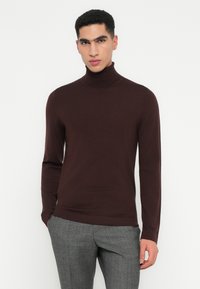 Bărbat îmbrăcat cu un pulover turtleneck dintr-o nuanță închisă de maro și pantaloni gri croiți, stând în fața unui fundal deschis simplu.