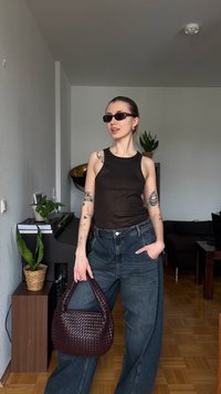 Νέα γυναίκα με γυαλιά ηλίου, μαύρη αμάνικη μπλούζα, φαρδιά τζιν, κρατώντας πλεκτή τσάντα, στέκεται σε ένα σαλόνι με φυτά και έπιπλα.