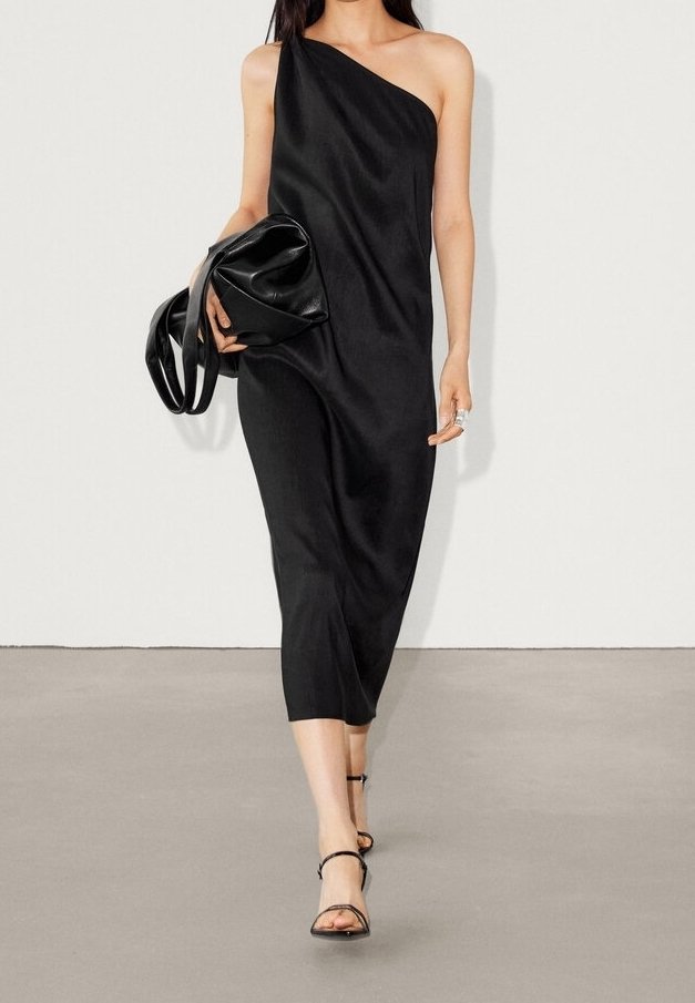 Robe noire asymétrique avec une silhouette fluide, longueur mi-mollet, et texture lisse. Associée à un sac à main noir arrondi et des sandales à lanières.