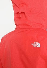Veste légère rouge avec texture verticale, dotée d'une capuche et d'un logo blanc à l'arrière. Détail avec cordon réglable au niveau du cou.