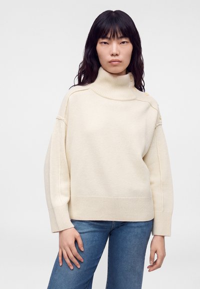 Femme aux longs cheveux noirs portant un pull à col roulé crème avec des boutons sur l'épaule et un jean bleu sur un fond uni.