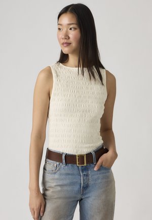 Femme aux longs cheveux bruns portant un haut blanc texturé sans manches, un jean bleu et une ceinture marron, regardant vers sa gauche avec la main dans la poche.