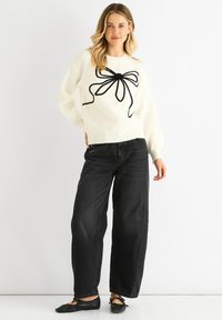 Vit stickad tröja med en svart rosett-design på framsidan, matchad med löst sittande svarta jeans och svarta platta skor. Slät textur.
