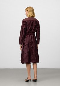 Kvinna med blont hår som bär en texturerad mörk burgunderröd midiklänning med bälte och svarta klacksandaler, står vänd mot en enkel ljusgrå vägg.