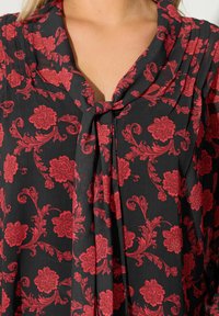 Sort bluse med et blomstermønster i røde og lyserøde nuancer, draperet halsudskæring og en bundet detalje i midten. Blødt, textured stof.