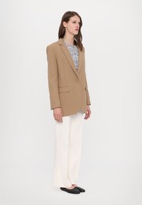 Beige villatakki, jossa on yksi nappi, viistetyt kaulukset, kaksi etutaskua, yhdistettynä valkoisiin leveälahkeisiin housuihin ja mustiin ballerinatossuihin.