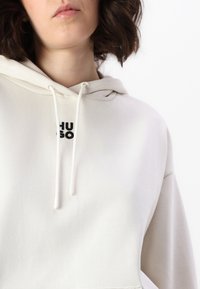 Světle béžová mikina s kapucí vyrobená z měkké látky. Obsahuje šňůrku na stažení kapuce a přední kapsu. Černé logo "HUGO" je výrazně zobrazeno na hrudi.