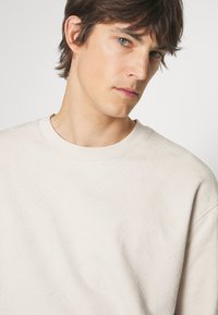 Beige sweatshirt met ronde hals en reliëf logo patroon, geribbelde halslijn, zachte textuur en een relaxte pasvorm. Subtiel ontwerp met minimalistische accenten.