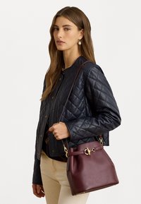 Granatowa pikowana skórzana kurtka, dopasowany krój, noszona na ciemnej bluzce. Burgundowa torba-bucket z złotymi akcentami, gładka faktura, pasek na ramię.