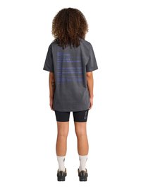 Persoon met krullend haar staat met de rug naar ons toe, draagt een loszittend donkergrijs T-shirt met blauwe tekst en zwarte fietsbroekjes met "ATW" op het linkerbeen.