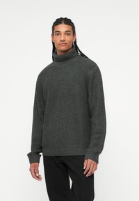 Mørkegrå ribbestrikket sweater med høj krave, struktureret strikkemønster og lange ærmer. Sat sammen med sorte bukser.