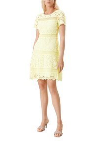 Nainen, jolla on lyhythihainen, polvenmittainen keltainen pitsimekko ja beigellä korkokengät, seisoo valkoista taustaa vasten.