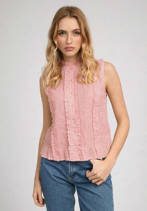 Femme blonde portant un chemisier rose à volants sans manches et un jean bleu, debout avec une main dans la poche devant un fond uni.