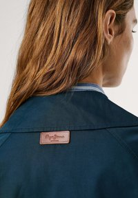 Veste bleu marine en tissu texturé, avec un patch en cuir au col arrière et un col intérieur à rayures contrastées.