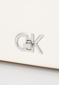 Bijela kožna torba s srebrnim metalnim kopčom i detaljem logotipa, s glatkom teksturom i minimalističkim dizajnom.