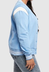 Person som bär en ljusblå varsityjacka med vita ränder på axlar, muddar och nederkant, tillsammans med urtvättade blåjeans, stående på sidan.