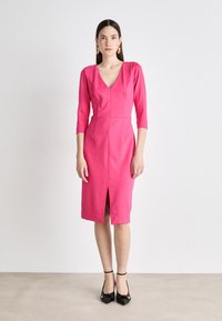 Fuchsia dress with a V-neck, three-quarter sleeves, front slit, and structured fit. Styled with black shoes featuring a knotted detail.