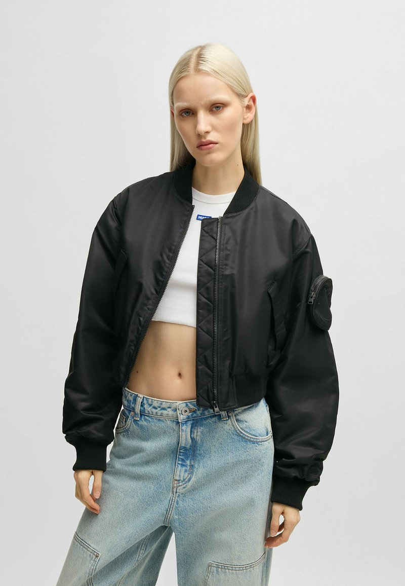 Svart bomberjacka med dragkedja, ribbade ärmslut och ett märke på ärmen. Bärs över en vit crop-top och ljusblå baggy-jeans.