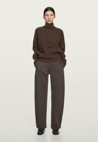 Brun ribbet rullekrave sweater parret med løstsiddende grå bukser. Modellen bærer en lang halskæde og ankelsko. Neutral baggrund.