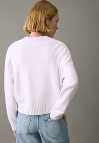 Bela pletena majica z rebrastim robom in manšetami. Ima sproščen kroj in spuščena ramena. Nošena z svetlo modrim kavbojkami iz jeansa.