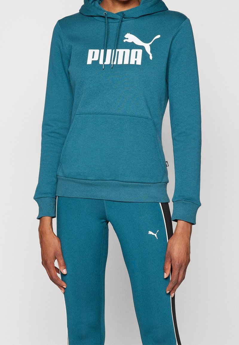 Personne portant un sweat à capuche Puma bleu sarcelle et un legging assorti avec des logos Puma blancs et des bandes latérales noires et blanches sur le legging.