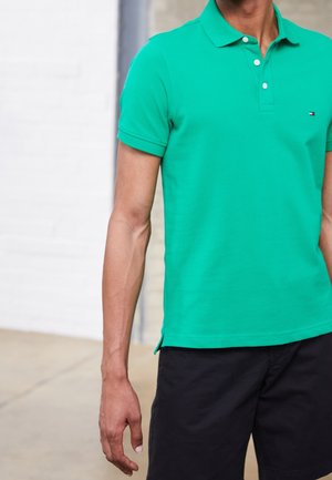 Homme portant un polo vert à manches courtes et un short noir, debout, les bras détendus le long du corps, devant un mur clair.