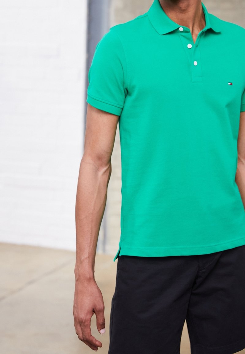 Homme portant un polo vert à manches courtes et un short noir, debout, les bras détendus le long du corps, devant un mur clair.