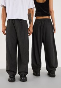 To personer iført løse mørkegrå bukser og sorte sko, den ene med en hvid kortærmet skjorte og tatoveret arm, den anden i en sort tanktop.