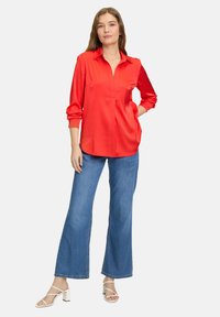 Rote Bluse mit langen Ärmeln, Kragen und vorderer Faltenlegung, kombiniert mit blauen weit geschnittenen Jeans und weißen Absatzsandalen.