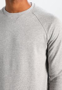 Sudadera gris con cuello redondo acanalado y mangas raglán, hecha de una tela suave. Textura lisa con un detalle de costura mínimo.