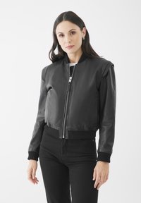 Černá kožená bomber bunda se zipem vpředu, pružnými manžetami a lemem, a hladkou texturou. Stylizováno s černými kalhotami a bílou košili pod ní.