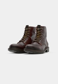 Botas de cuero marrón hasta el tobillo con puntas redondas, superficie texturizada y ojales de metal. Presentan cordones marrones y grises y suelas de goma negra resistentes.