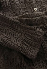 Sötétbarna, texturált szövet, ráncolt, gumis részletekkel és egy kis fehér gombbal a tetején, középen.