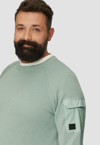 Lichtgroene waffle-gebreide sweatshirt met een ronde halslijn, geribbelde manchetten en een patch pocket op de linkermouw met een zwarte accenten.