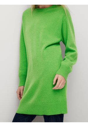 Femme portant une robe-pull à manches longues vert vif avec poignets et ourlet côtelés, associée à un pantalon foncé.