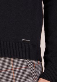 Pull en laine bleu marine avec ourlet et poignets côtelés, arborant une petite étiquette de marque en argent. Associé à un pantalon à carreaux gris et noir.