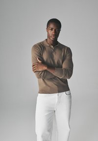 Braunes, langärmliges Polohemd mit Kragen und gerippten Bündchen, kombiniert mit weißen Hosen. Einfaches Design, glatte Textur und keine sichtbaren Logos.