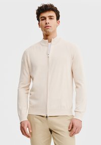 Jasno beżowy sweter na zamek wykonany z miękkiego materiału, z kołnierzem stojącym i prążkowanymi mankietami, w połączeniu z brązowymi spodniami.